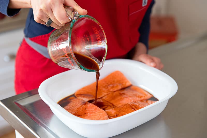 Pouring All-Purpose Stir-Fry Sauce from a measuring cup onto four salmon fillets in a white square container.