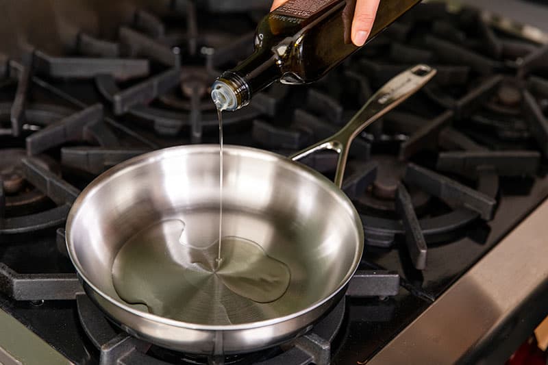 An overhead shot of someone pouring extra virgin olive oil into a hot stainless steel skillet.