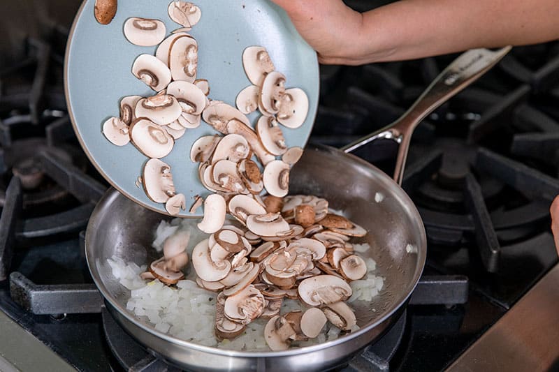 Adding a plate of thinly sliced cremini mushrooms to a skillet filled with diced onions.