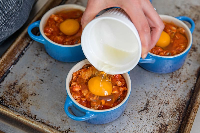 Someone pouring in a raw egg into a blue cocotte filled with spicy tomato meat sauce
