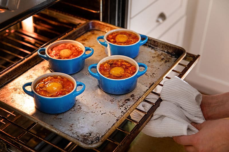 A rimmed baking sheet with four blue cocottes filled with eggs in purgatory are being placed into a hot oven.