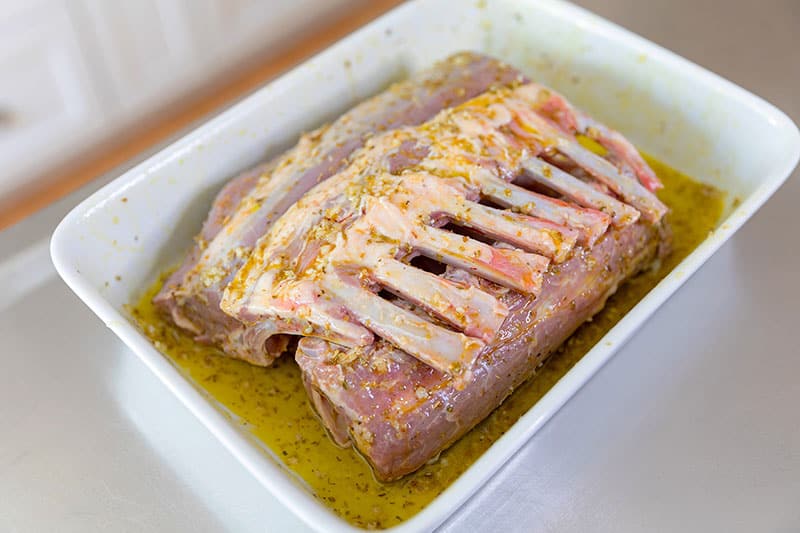 A white rectangular baking dish is filled with two lamb racks in a garlic lemon marinade.