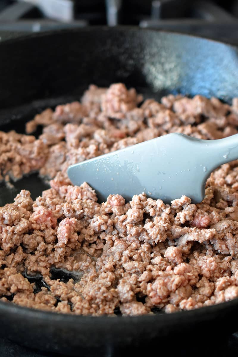 Frying ground beef in a cast iron skillet and stirring it with a gray silicone spatula. The beef is fully cooked and no longer pink.