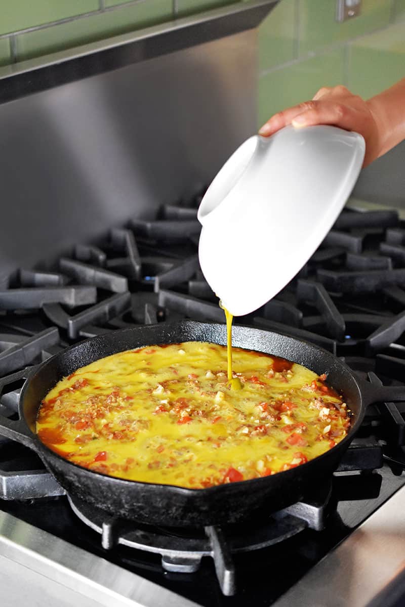 Pouring whisked eggs into a cast iron skillet filled with ingredients to make Whole30 and keto Tex Mex Beef and Rice Casserole.