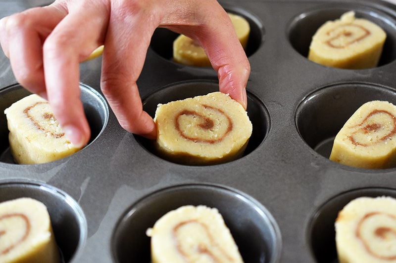 Placing sliced cinnamon rolls, flat-side down, into a greased muffin tin well. 