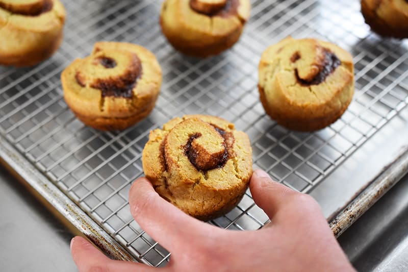A hand is placing paleo cinnamon rolls on a wire rack in a rimmed baking sheet.