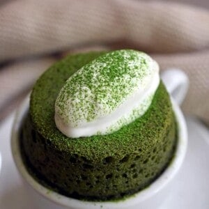 A close-up of a grain-free and gltuen-free matcha mug cake puffed over a white coffee mug and topped with dairy-free coconut whipped cream.