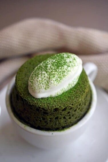 A close-up of a grain-free and gltuen-free matcha mug cake puffed over a white coffee mug and topped with dairy-free coconut whipped cream.