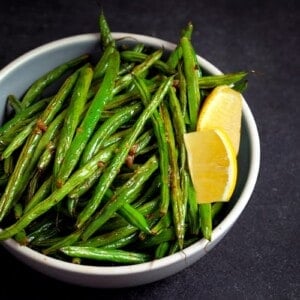 A bowl filled with roasted green beans with lemon and garlic and two lemon slices.