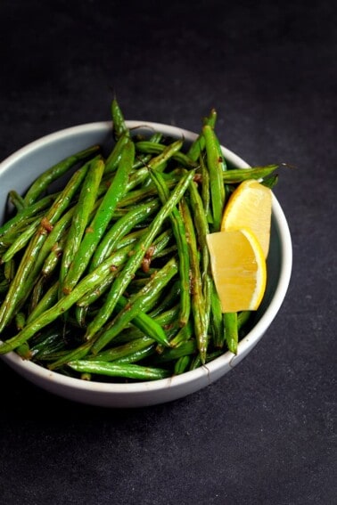 A bowl filled with roasted green beans with lemon and garlic and two lemon slices.