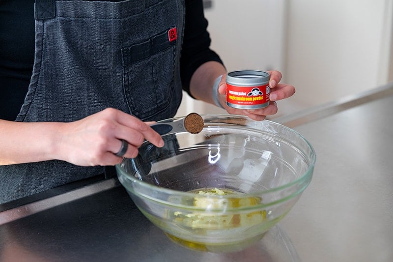 Measuring the marinade ingredients for roasted green beans with lemon and garlic into a large glass mixing bowl. A woman is adding a spoonful of Nom Nom Paleo's Magic Mushroom Powder to the bowl.