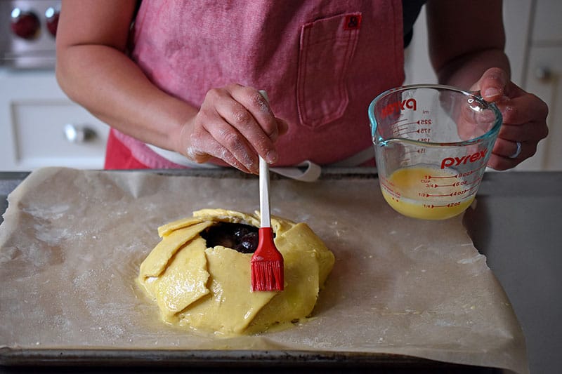 Someone using a red silicone brush to dab egg wash onto the cherry galette dough.