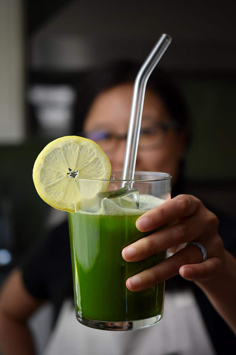 A woman in glasses is holding up a glass of matcha lemonade