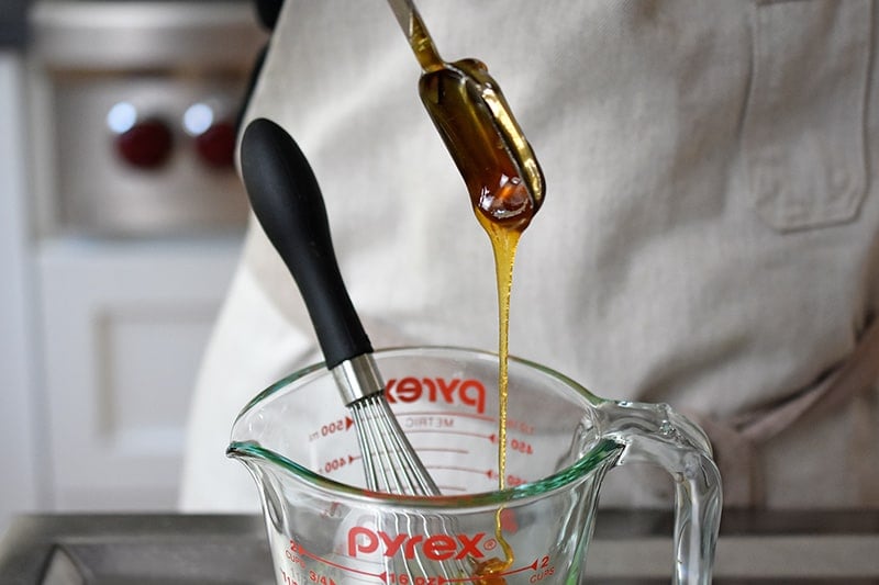 Adding a spoonful of honey to a liquid measuring cup with a small hand whisk inside.