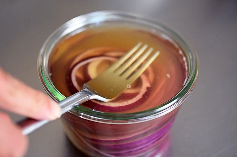 A fork pressing red onion slices in the brine to make Whole30 quick pickled red onions.