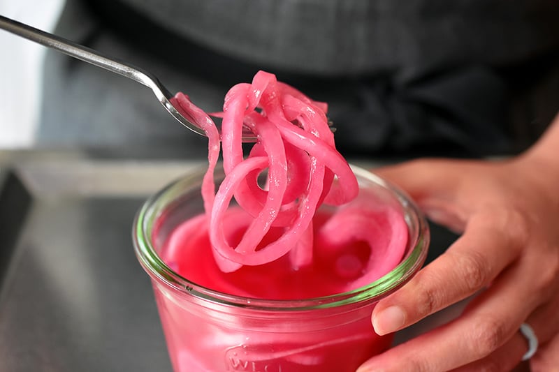 A fork pulling out a tangle of quick pickled red onions from a clear glass jar.