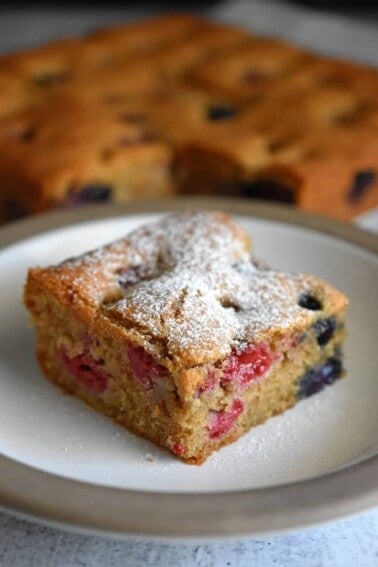 A slice of paleo and gluten-free berry snack cake is on a white plate. In the background are the other slices of berry snack cake.