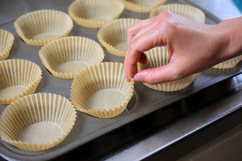 A hand is lining a muffin tin with parchment cupcake liners.