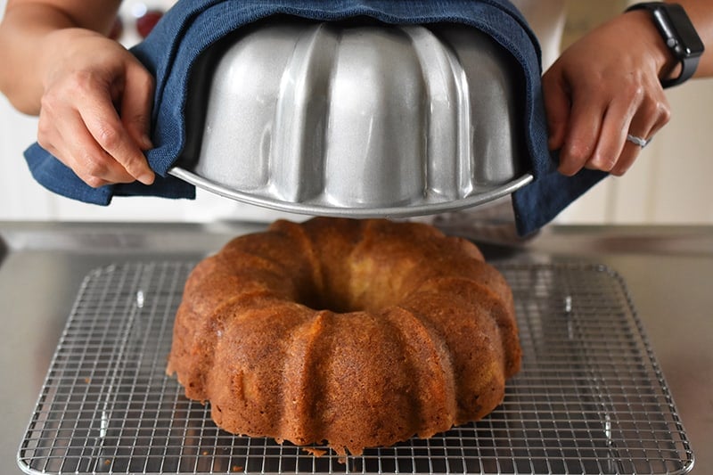 Inverting the apple Bundt Cake on a wire rack and removing the pan to reveal a golden brown cake.