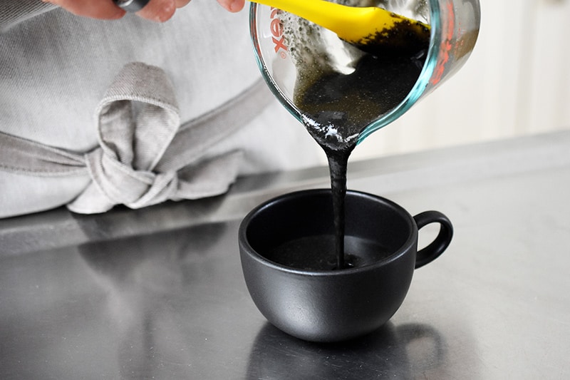 Pouring the black sesame mug cake batter into a black tea cup.