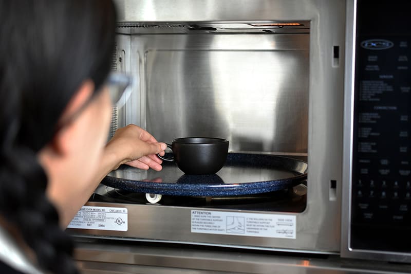 A brunette woman with glasses placing a black tea cup into a microwave.