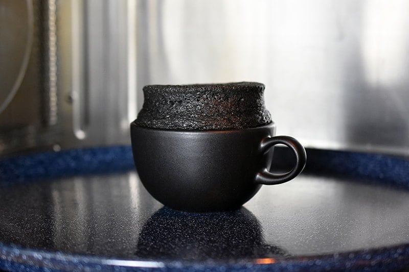Looking in a microwave at a black tea cup filled with black sesame cake that's puffed up and ready to eat.