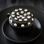 A black mug on a two black plates is filled with a black sesame cake topped with candy eyeballs.
