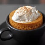 A black coffee mug filled with paleo and gluten-free pumpkin mug cake cooked in the microwave.The cake is topped with whipped coconut cream and dusted with pumpkin spice blend.
