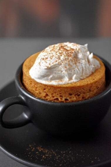 A black coffee mug filled with paleo and gluten-free pumpkin mug cake cooked in the microwave.The cake is topped with whipped coconut cream and dusted with pumpkin spice blend.