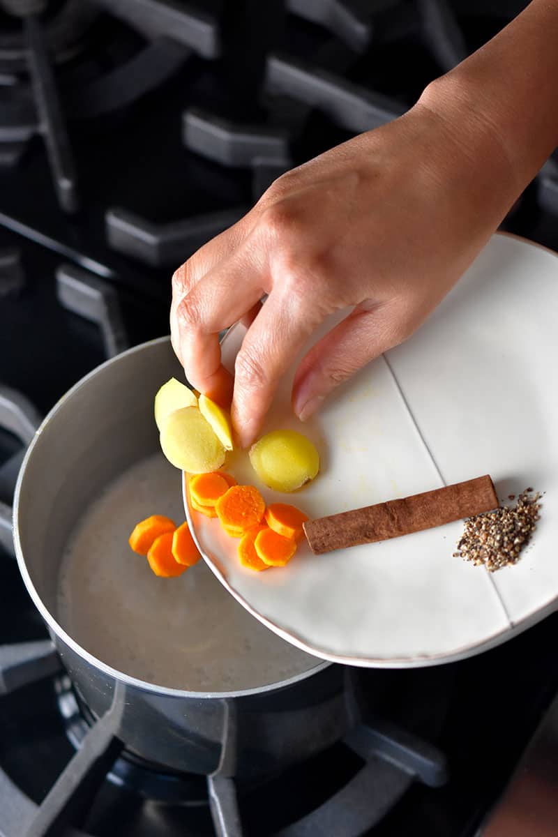 A hand adding turmeric slices, ginger slices, a cinnamon stick, and ground pepper to a saucepan filed with coconut milk to make golden milk.