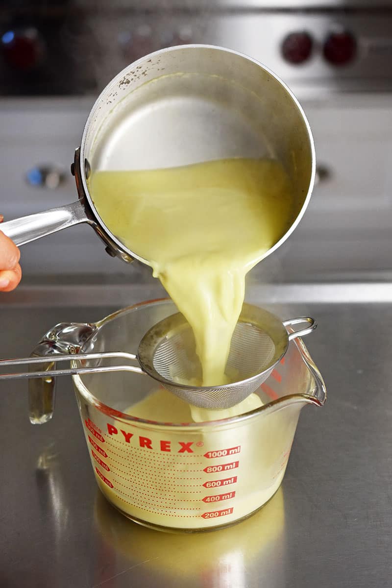 Pouring the Golden Milk from a small saucepan through a fine mesh sieve into a 4-cup Pyrex liquid measuring cup.