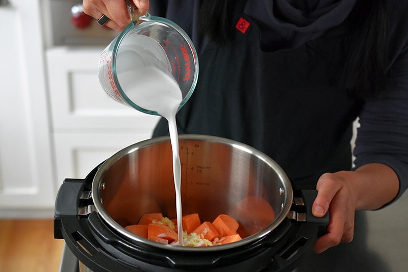 Pouring coconut milk into an Instant Pot filled with cubed sweet potatoes and minced garlic.