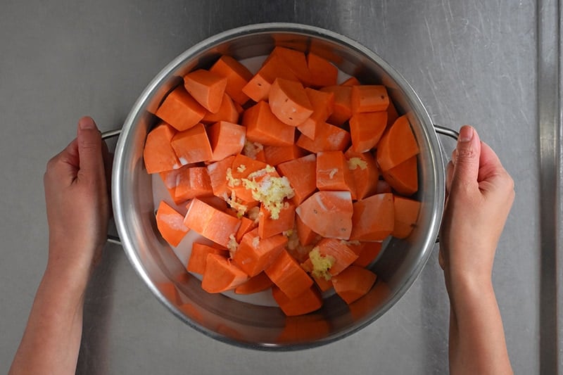 An overhead shot of two hands holding a pot with cubed sweet potatoes, coconut milk, garlic, and salt.