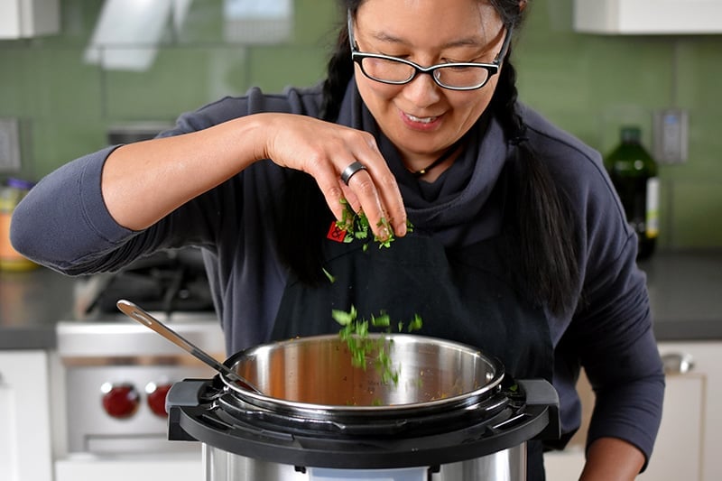A brunette smiling woman is adding minced cilantro to an open Instant Pot