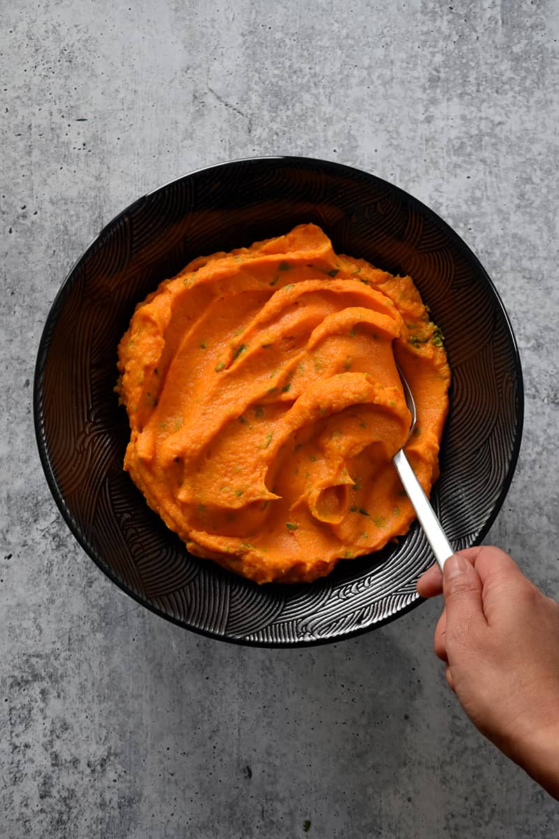 An overhead shot of a a hand scooping vegan and paleo Instant Pot Mashed Sweet Potatoes from a black bowl.