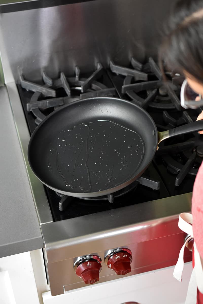 A 12-inch non-stick skillet is being tipped on a gas stovetop to swirl the oil inside.