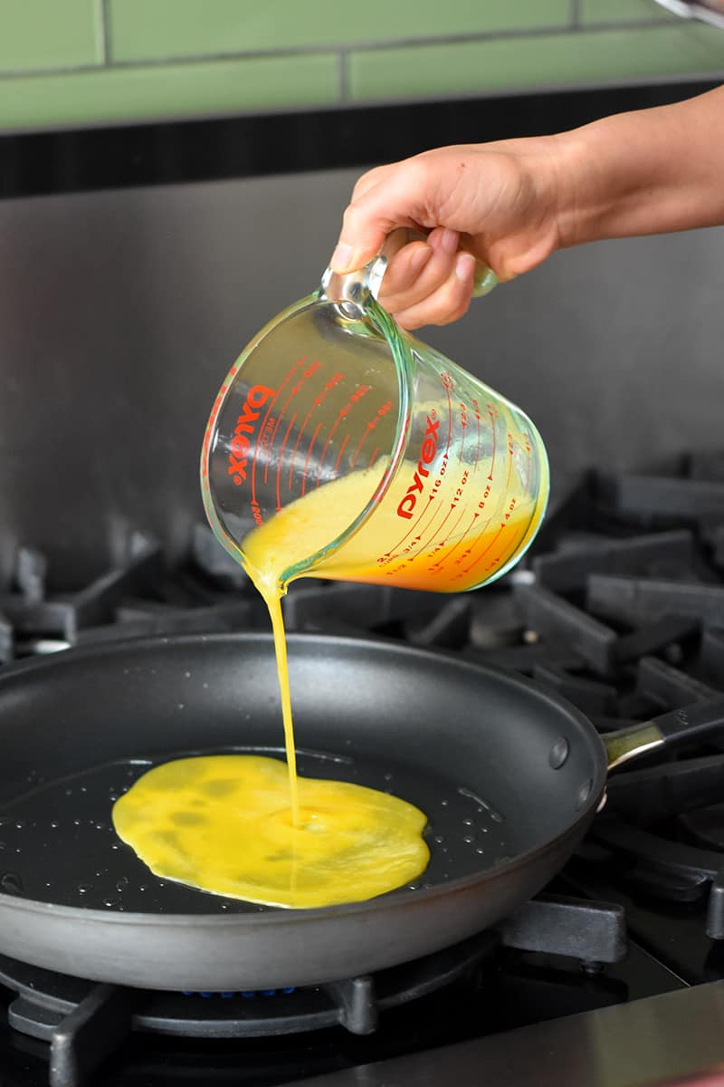 A hand pouring whisked eggs from a Pyrex measuring cup into a 12-inch Oxo non-stick skillet.