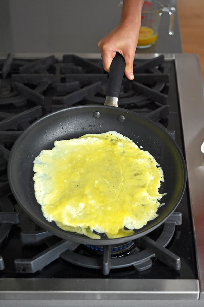 A hand is tipping a large non-stick skillet to make a thin layer of egg omelet on the bottom of the skillet.