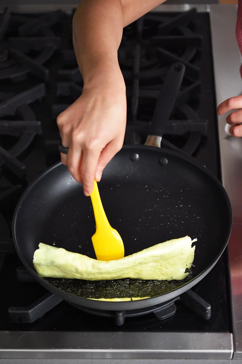A hand is using a yellow silicone spatula flipping a Korean egg roll with roasted seaweed. The Korean egg roll is almost flipped all the way at the end.
