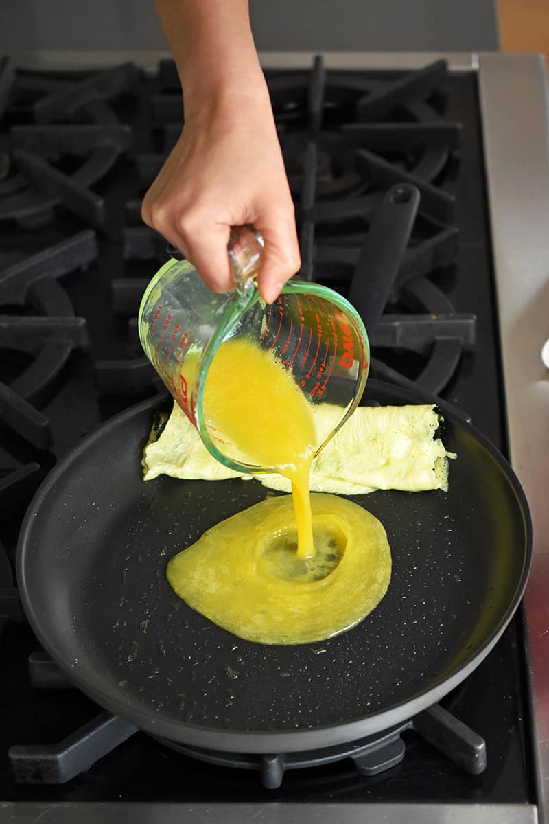 A hand pours whisked eggs from a measuring cup into a non-stick skillet that has a cooked egg roll omelet on the edge.