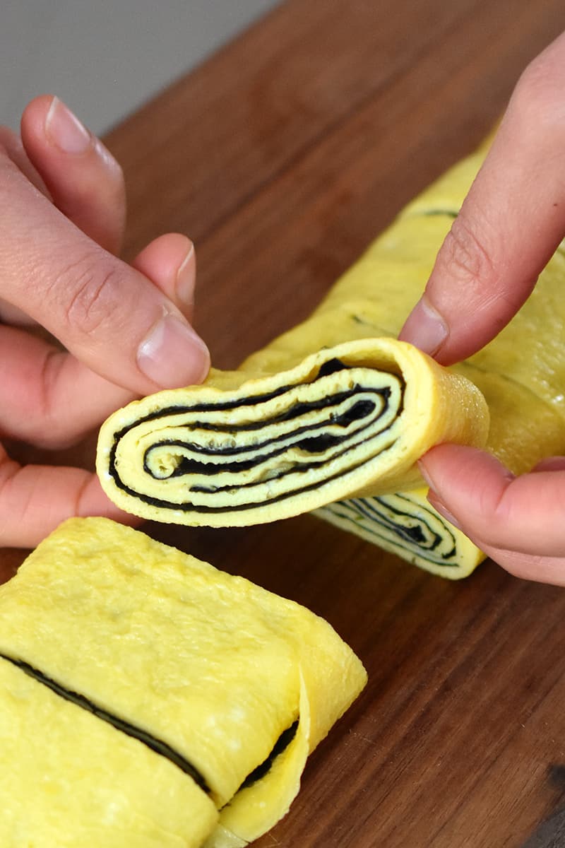 Two hands are holding up a slice of Korean rolled omelet with toasted seaweed folded inside.