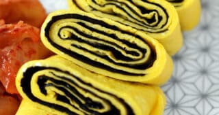 A closeup shot of a cut up Korean Egg Roll Omelet with Roasted Seaweed on a white plate with gray flowers. You can see the spirals of egg and bright yellow egg in the cut-up pieces.
