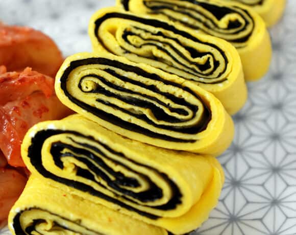 A closeup shot of a cut up Korean Egg Roll Omelet with Roasted Seaweed on a white plate with gray flowers. You can see the spirals of egg and bright yellow egg in the cut-up pieces.