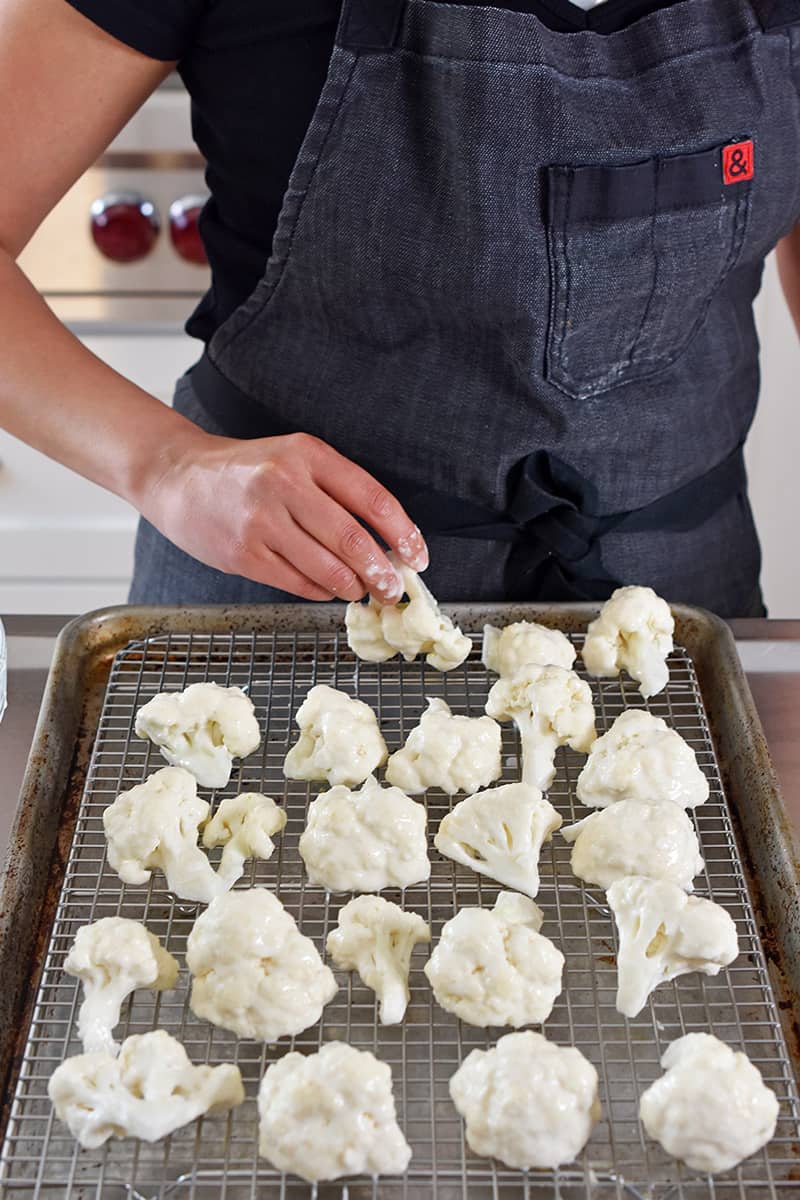 Someone in a gray apron is placing battered cauliflower on a wire rack in a rimmed baking sheet.