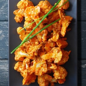 An overhead shot of baked buffalo cauliflower on a rectangular gray plate. There are three chives on top.