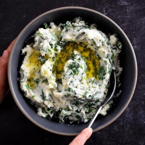A hand is scooping paleo cauliflower colcannon from a gray bowl. The colcannon is white pureed cauliflower with flecks of green kale and scallions in it. There is melted ghee on top.