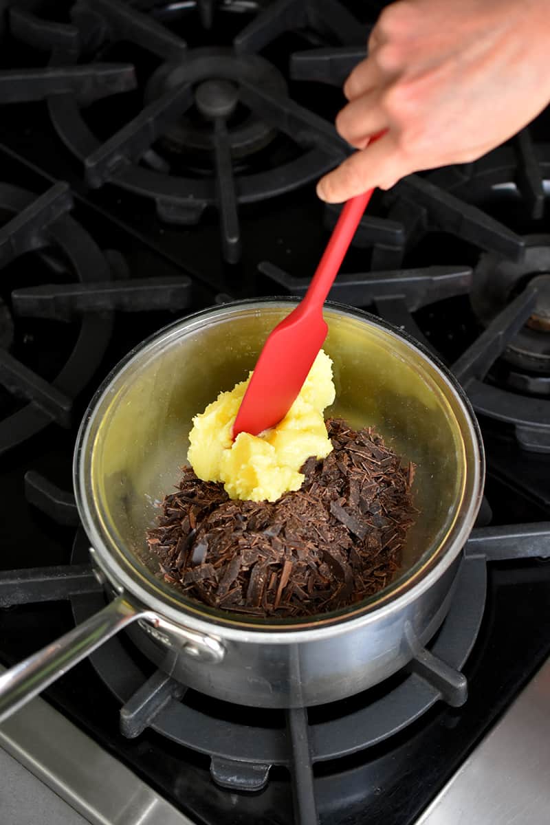 A red silicone spatula is adding ghee to a bowl filled with chocolate shards. The bowl is on top of a large saucepan.