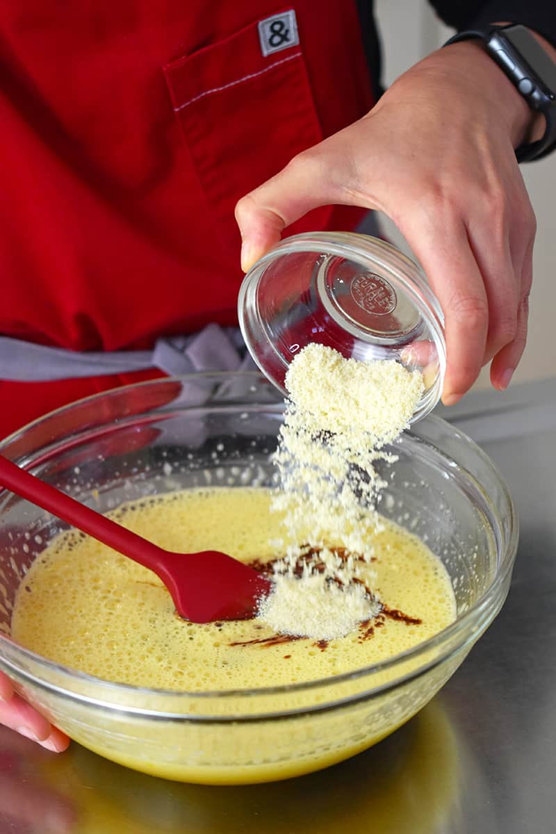 Someone adding almond flour from a small glass bowl into a large bowl filled with lava cake batter.