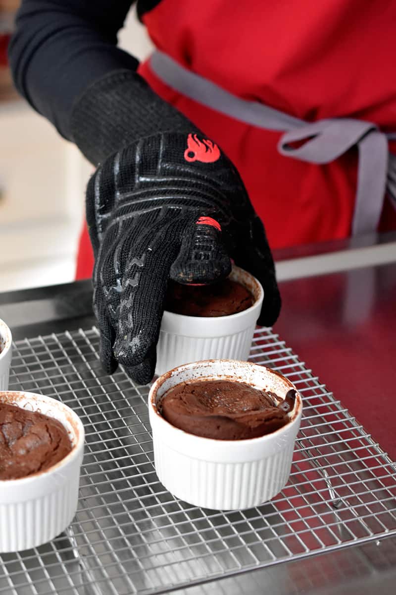 A hand in a kevlar glove is transferring cooked lava cakes onto a wire rack.