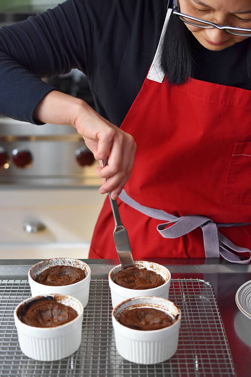 A woman in a red apron is using a butter knife to cut around the edge of a lava cake. 